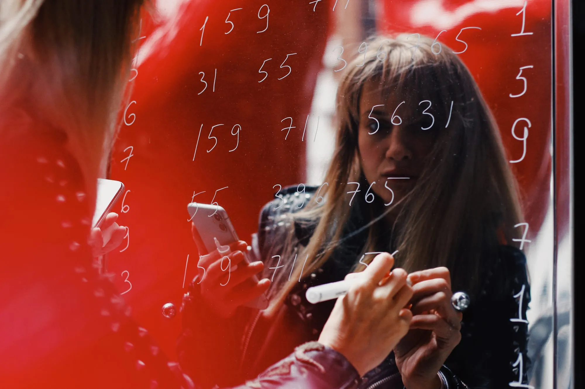 Asya Aseeva writing numbers on a glass panel, her reflection doubled against a red surface